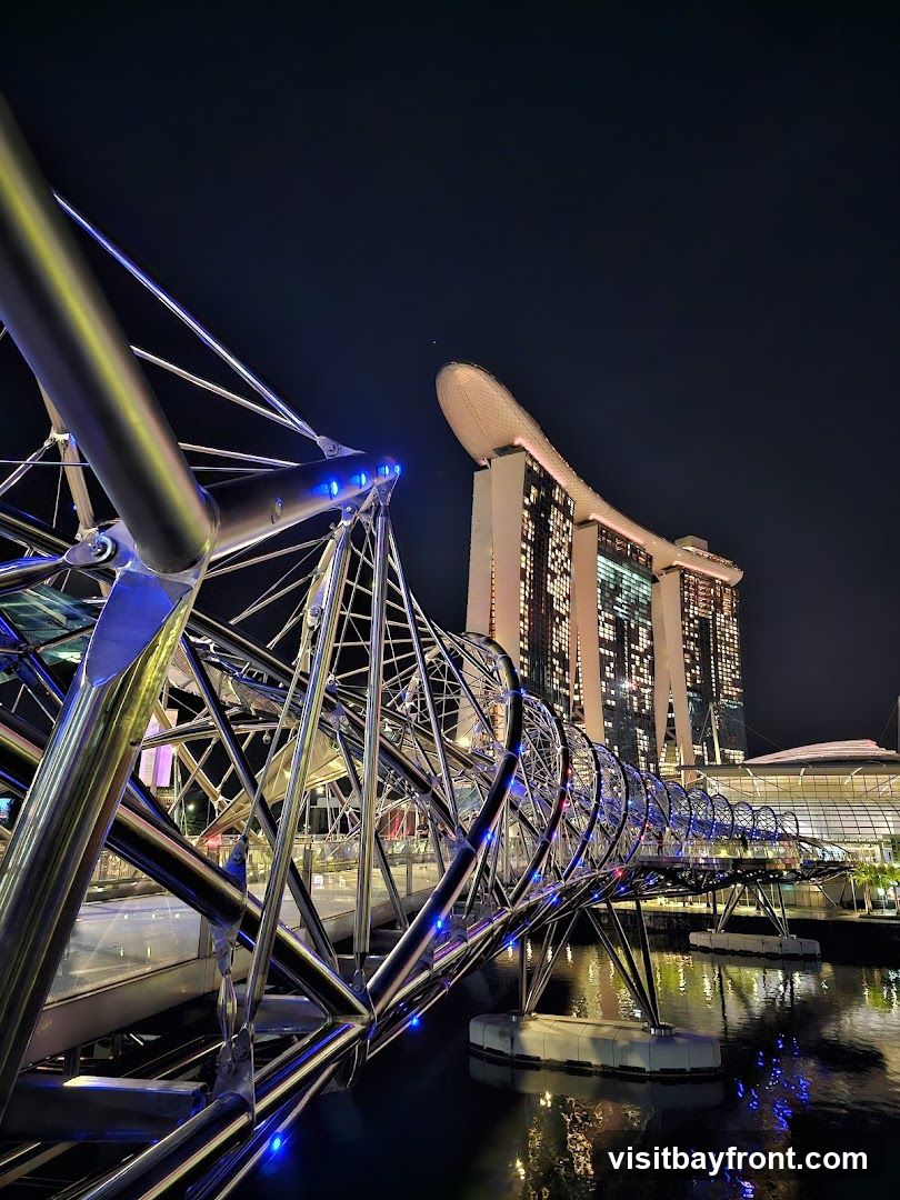 Night view of the Helix Bridge in Singapore with illuminated blue lights and Marina Bay Sands hotel in background.
