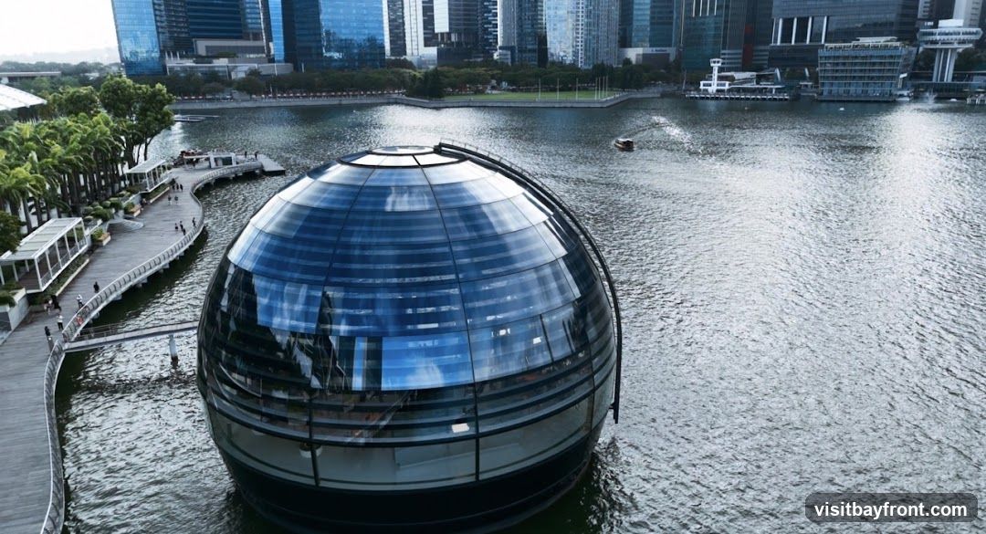 Aerial view of the Sphera, a spherical glass building on Marina Bay in Singapore. The structure is surrounded by water with modern skyscrapers and waterfront promenades visible in the background.