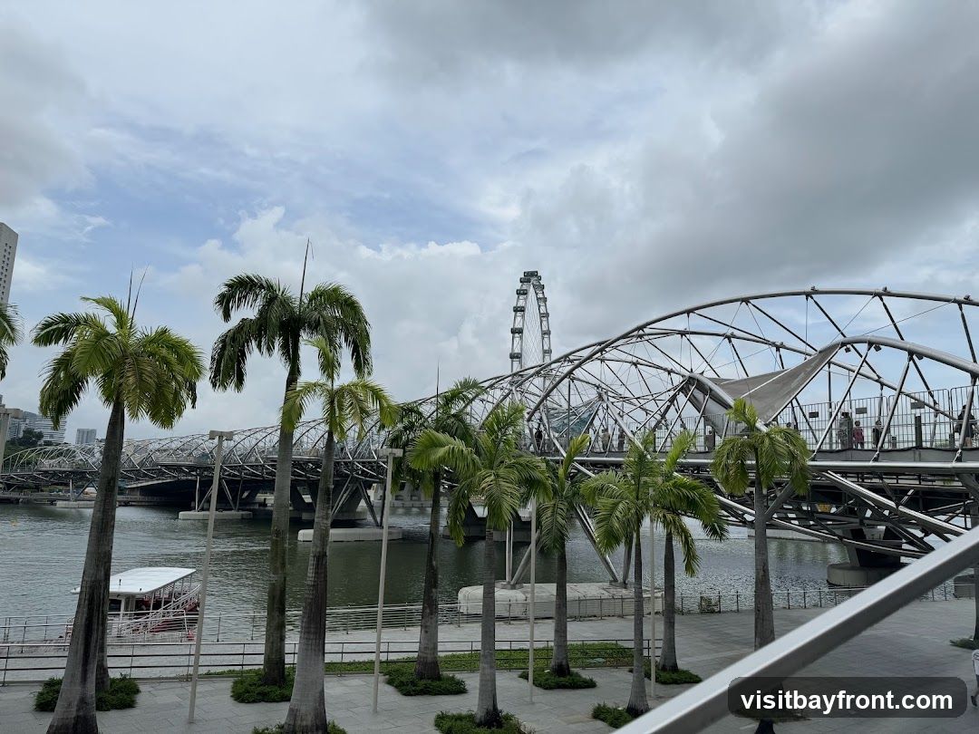The image shows a modern residential building (likely an HDB block in Singapore) viewed from across a river or canal. In the foreground, there are several palm trees lining a paved walkway. A distinctive arched pedestrian bridge spans the water, connecting to the area near the building. In the background, a Ferris wheel is visible against a cloudy sky. The scene appears calm and urban, with no visible shop fronts, menus, or commercial signage.