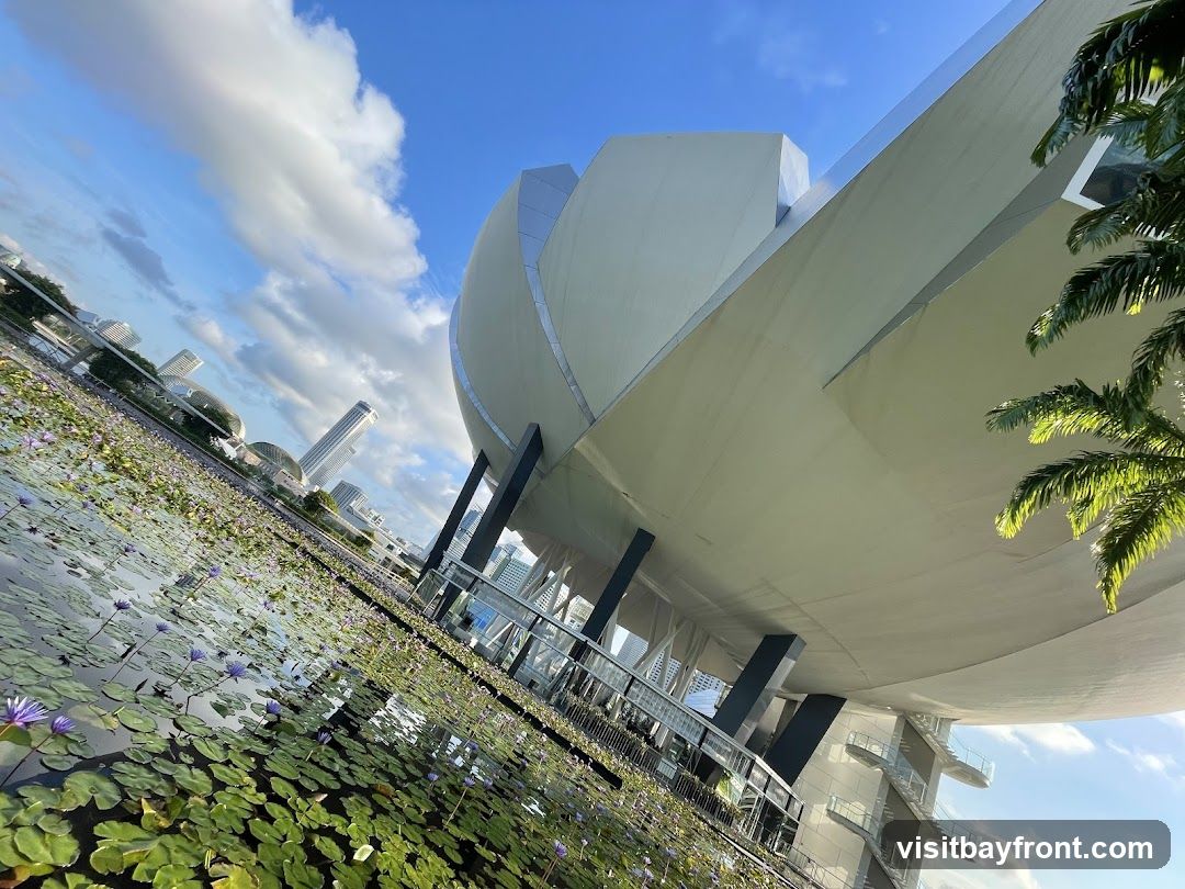 A low-angle view of a modern white building resembling the Esplanade – Theatres on the Bay in Singapore, featuring curved architectural lines and large overhanging roof structures supported by dark columns. In the foreground, a pond filled with green lily pads and purple water lilies stretches across the bottom left. Palm fronds frame the right side. The sky is bright blue with scattered white clouds. Distant city skyscrapers are visible in the background under daylight conditions.