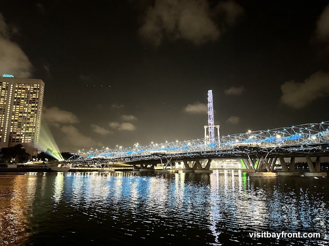 A nighttime scene featuring the illuminated Helix Bridge spanning across a body of water, with reflections shimmering on the surface. To the left, a tall building is lit up, while in the background, a distinctive tower structure stands against the dark sky dotted with clouds.