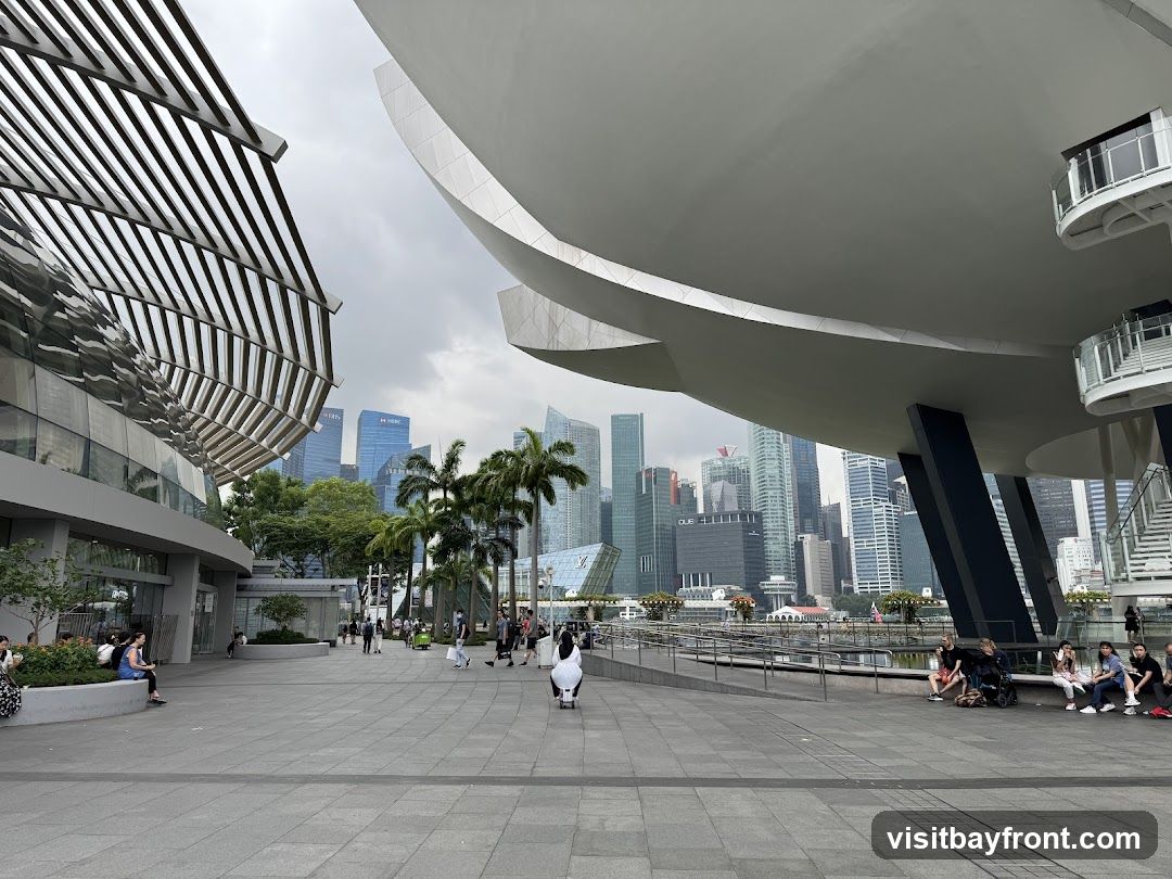 A wide-angle view of the ArtScience Museum in Singapore, with its distinctive lotus-shaped architecture dominating the foreground. The museum's white, curved structure contrasts with the modern glass buildings of the financial district in the background. People are seen walking along the promenade, enjoying the urban waterfront setting.