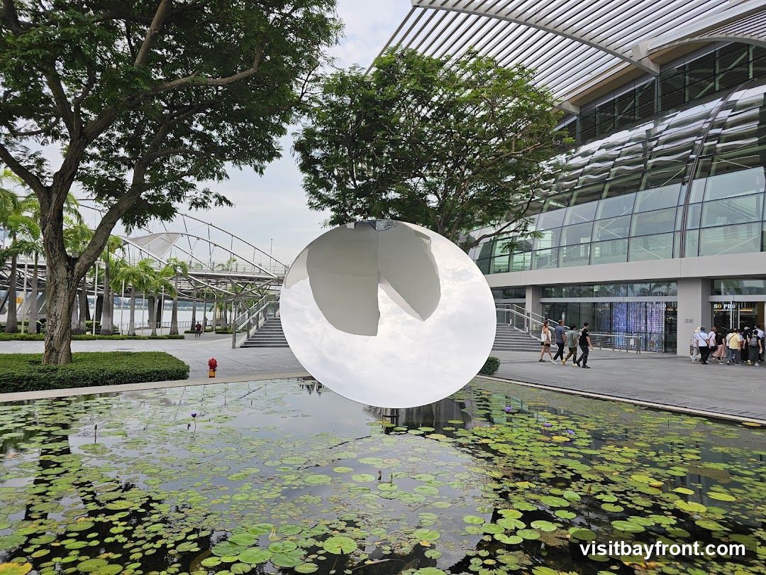 A large white spherical sculpture floats above a lily pad-covered pond in front of the modern glass facade of Changi Airport Terminal 3. People walk along the paved plaza near the entrance, and palm trees frame the scene under an overcast sky.