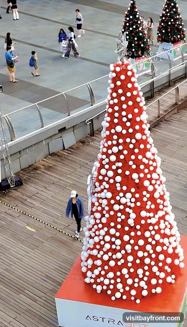 A large red Christmas tree decorated with white pom-poms stands on a wooden platform labeled 'ASTRA301'. In the background, people walk along a paved area near a railing, and additional smaller Christmas trees are visible.