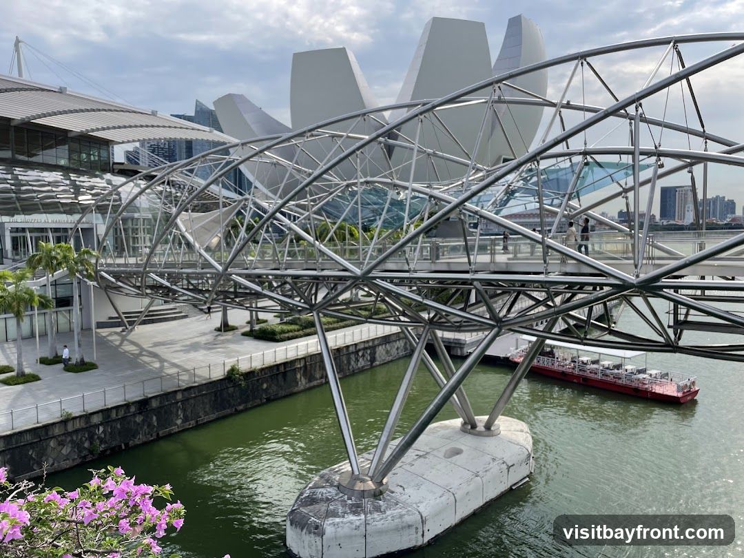The image shows the Helix Bridge in Singapore, a distinctive double-decker pedestrian bridge with its unique helical design. In the background, the ArtScience Museum is visible with its lotus-like architecture. A red boat is passing under the bridge on the water. The scene includes palm trees and flowering plants along the walkway.