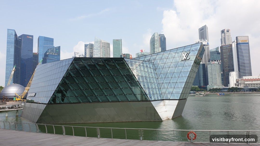 A modern glass building with a distinctive angular design, featuring the Louis Vuitmon logo on its facade. The structure is situated near water with a city skyline in the background.