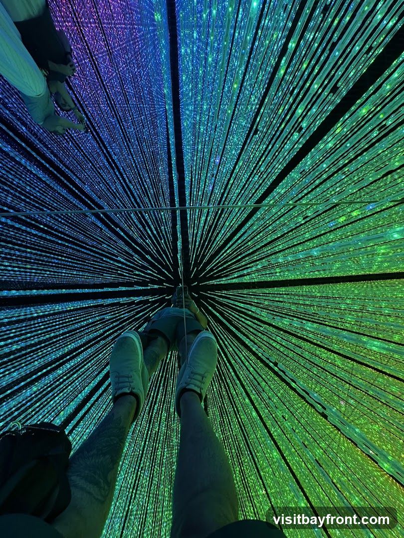 First-person view looking up at a ceiling installation of thousands of vertical LED strands radiating outward from the center, creating a starburst effect with blue and green hues. The viewer's legs and feet are visible in the foreground.