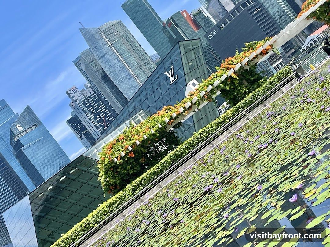A vibrant cityscape featuring a modern glass building adorned with lush greenery and colorful flowers along its facade. In the foreground, a serene water feature dotted with lily pads and purple water lilies adds tranquility to the bustling urban environment. The background showcases towering skyscrapers under a clear blue sky, highlighting the blend of nature and architecture in this metropolitan setting.