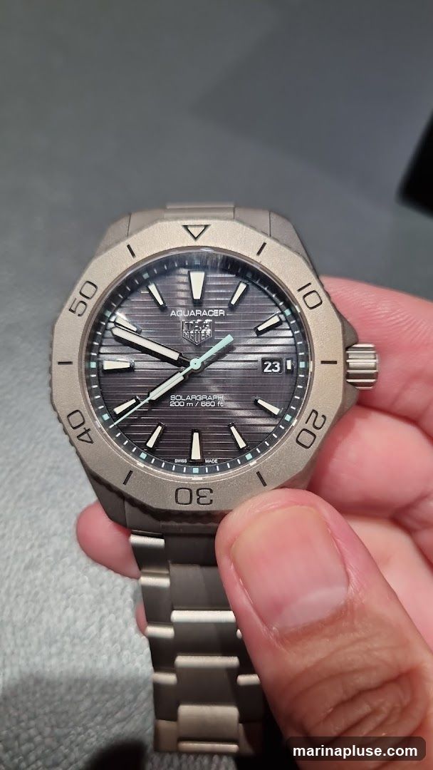 A close-up shot of a TAG Heuer Aquaracer Solargraph watch being held by a person. The watch has a dark, textured dial with silver hour markers and hands. The bezel is gray and has minute markers. The watch band is also gray and made of metal links. The date window shows '23'.