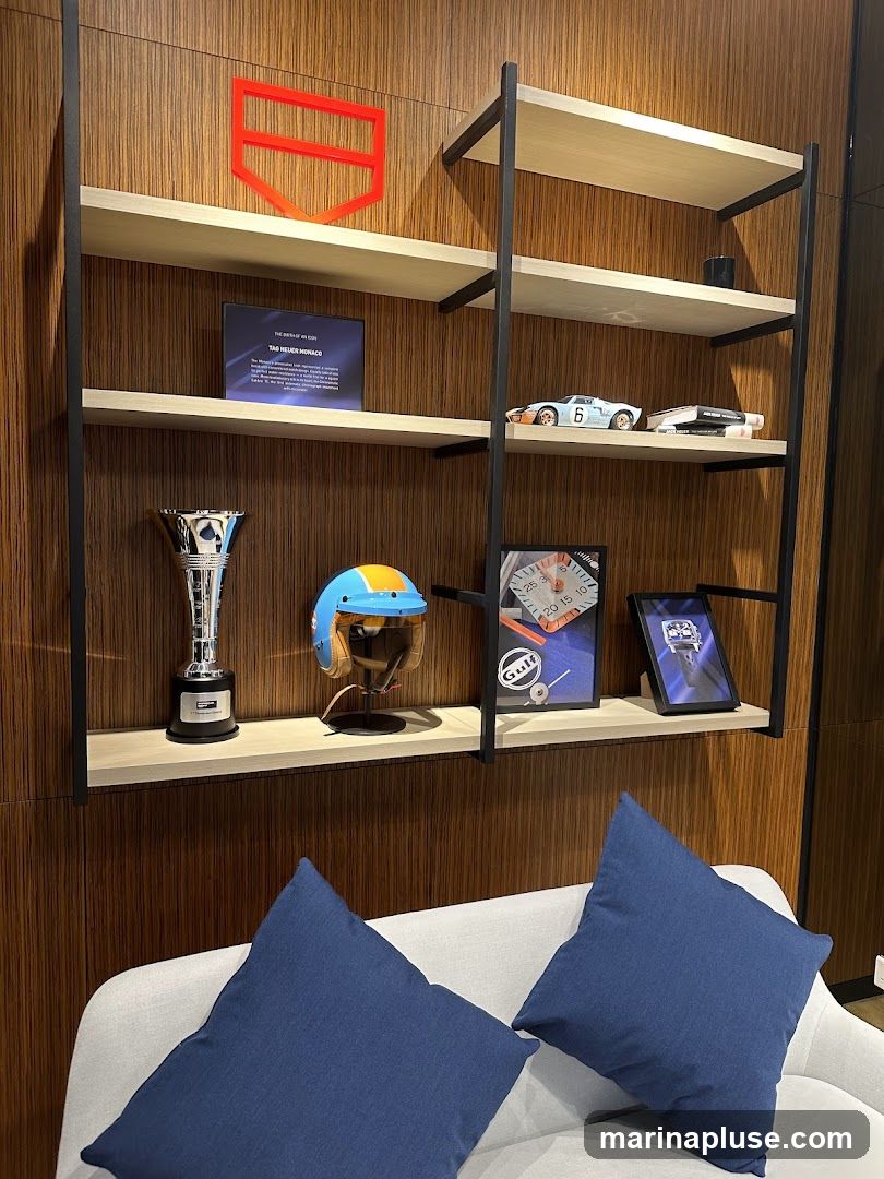 A wall-mounted shelving unit displays various items related to racing and watches. The top shelf features a red, abstract logo. The second shelf holds a framed plaque with text and a model race car. The third shelf displays a trophy, a racing helmet, and two framed pictures, one of which appears to be a watch face. The shelves are set against a wood-paneled wall, and a sofa with blue cushions is visible in the foreground.