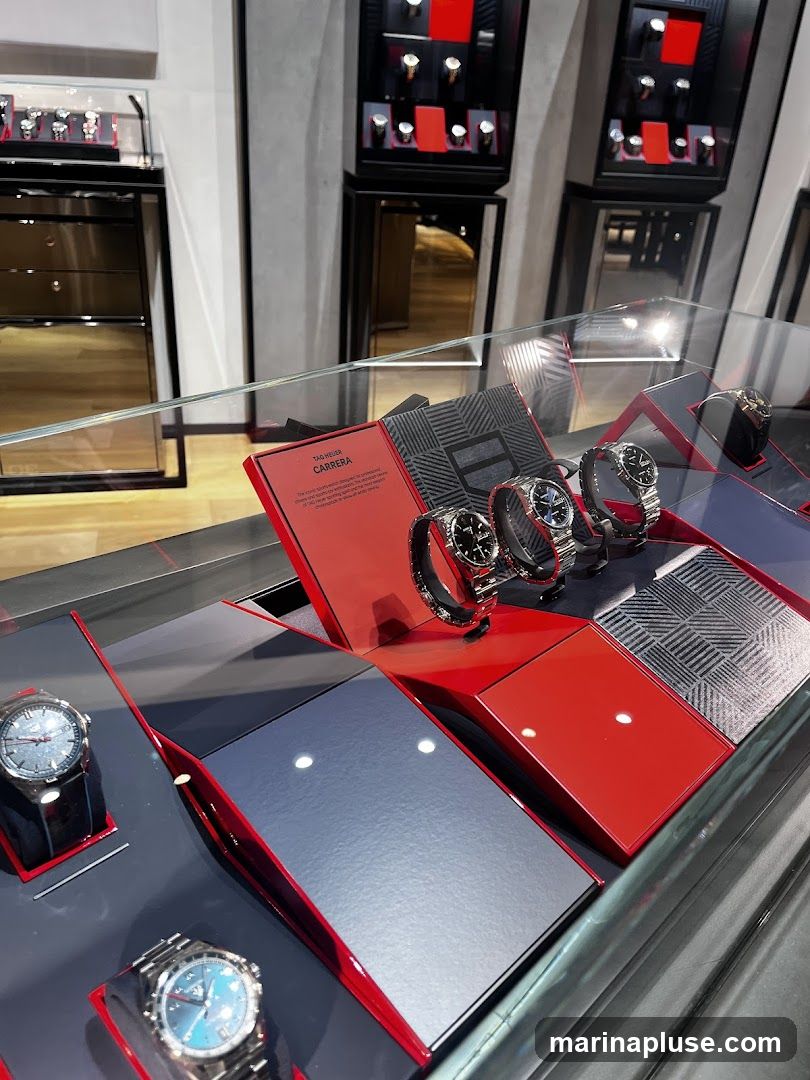 A display case filled with various watches. In the foreground, three watches are visible on stands. Behind them, a red box with the text "TAG HEUER CARRERA" is open, showcasing several watches. The background shows more watch displays in cabinets.