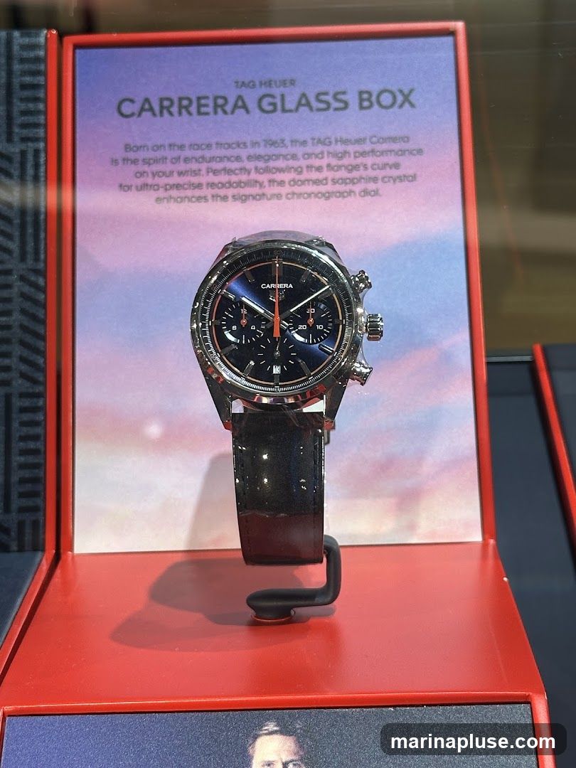 A TAG Heuer Carrera watch is displayed in a red and purple stand. The watch has a black leather strap and a dark blue dial with orange accents. A descriptive text panel above the watch reads: "TAG HEUER CARRERA GLASS BOX Born on the race tracks in 1963, the TAG Heuer Carrera Is the spirit of endurance, elegance, and high performance on your wrist. Perfectly following the flange's curve for ultra-precise readability, the domed sapphire crystal enhances the signature chronograph dial."