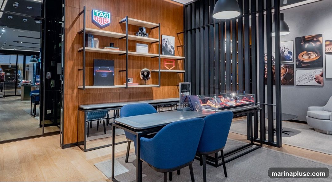 The interior of a modern TAG Heuer store features a wooden paneled wall with shelves displaying products and artwork. A central display table with blue chairs is set up for customer consultations. A glass display case showcases watches and accessories. The store has a clean and luxurious aesthetic with ample lighting.