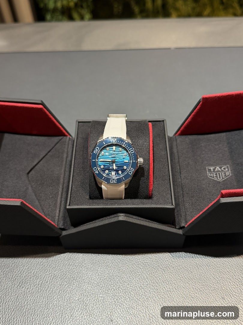 A close-up shot of a TAG Heuer Aquaracer watch with a blue wave-patterned dial and a white rubber strap, displayed in its open black and red presentation box. The TAG Heuer logo is visible on the inside lid of the box.