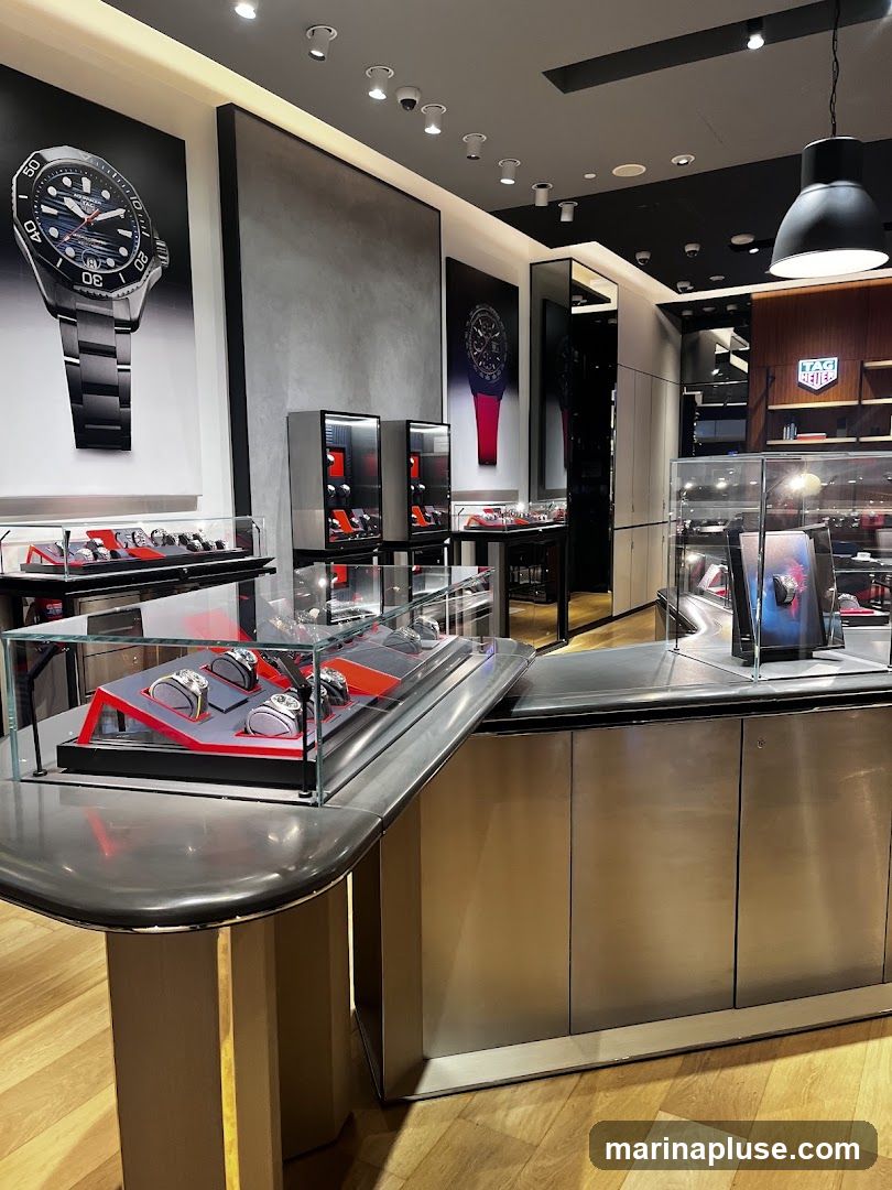 The interior of a modern watch store, featuring display cases filled with watches. Large posters of watches are mounted on the walls. The store has a sleek, metallic design with wooden flooring. A prominent TAG Heuer logo is visible on a shelf in the background.