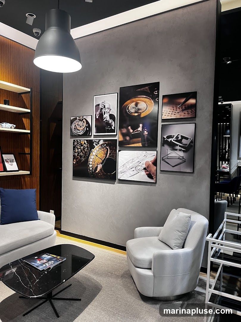 A modern lounge area with two plush armchairs, a black marble coffee table, and a grey rug. The walls are adorned with framed artwork, featuring close-ups of intricate mechanical components and a portrait of a photographer. A large pendant light hangs from the ceiling. To the left, a wooden shelf displays a model car and framed pictures. To the right, a white shelving unit is visible.