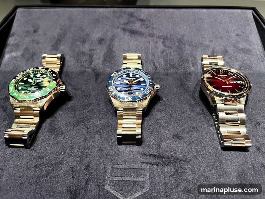 Three luxury watches with metal bands are displayed on a dark grey, textured surface. The watch on the left has a green bezel and a green dial. The middle watch has a blue bezel and a blue dial with orange accents. The watch on the right has a red dial and a silver bezel. All watches appear to be automatic.
