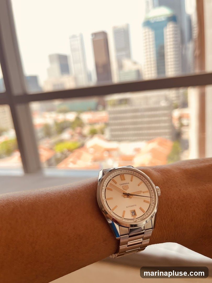 A close-up shot of a person's wrist wearing a silver watch with a cream-colored face. The watch has the brand name "CARRERA" and "TAG HEUER" visible. The background is a blurred view of a city skyline with tall buildings and some lower-rise structures.