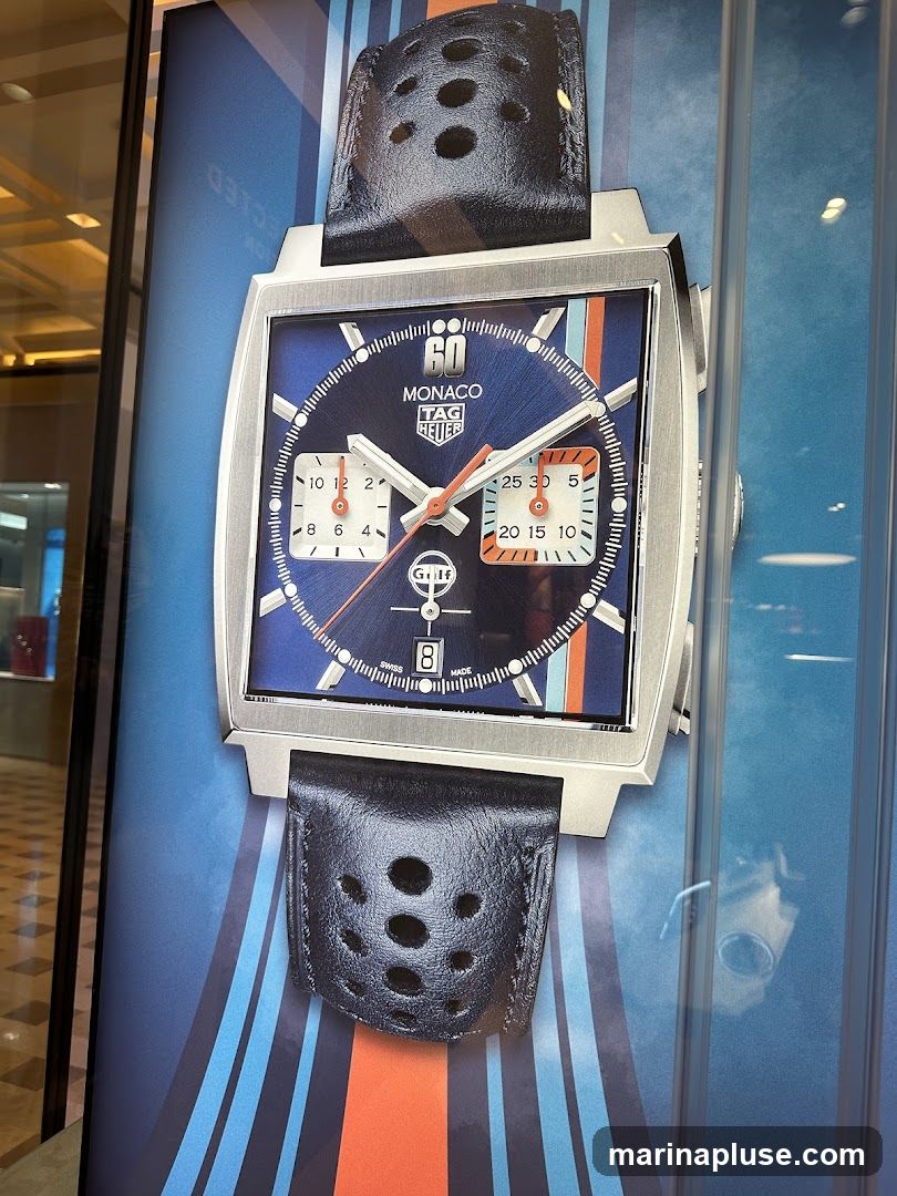 A close-up image of a TAG Heuer Monaco watch displayed on a blue background with racing stripes. The watch features a square case, a blue dial with white and orange accents, and a black leather strap with perforations. The dial shows the "TAG Heuer" logo, "MONACO" text, and sub-dials. The background has vertical blue and orange stripes.