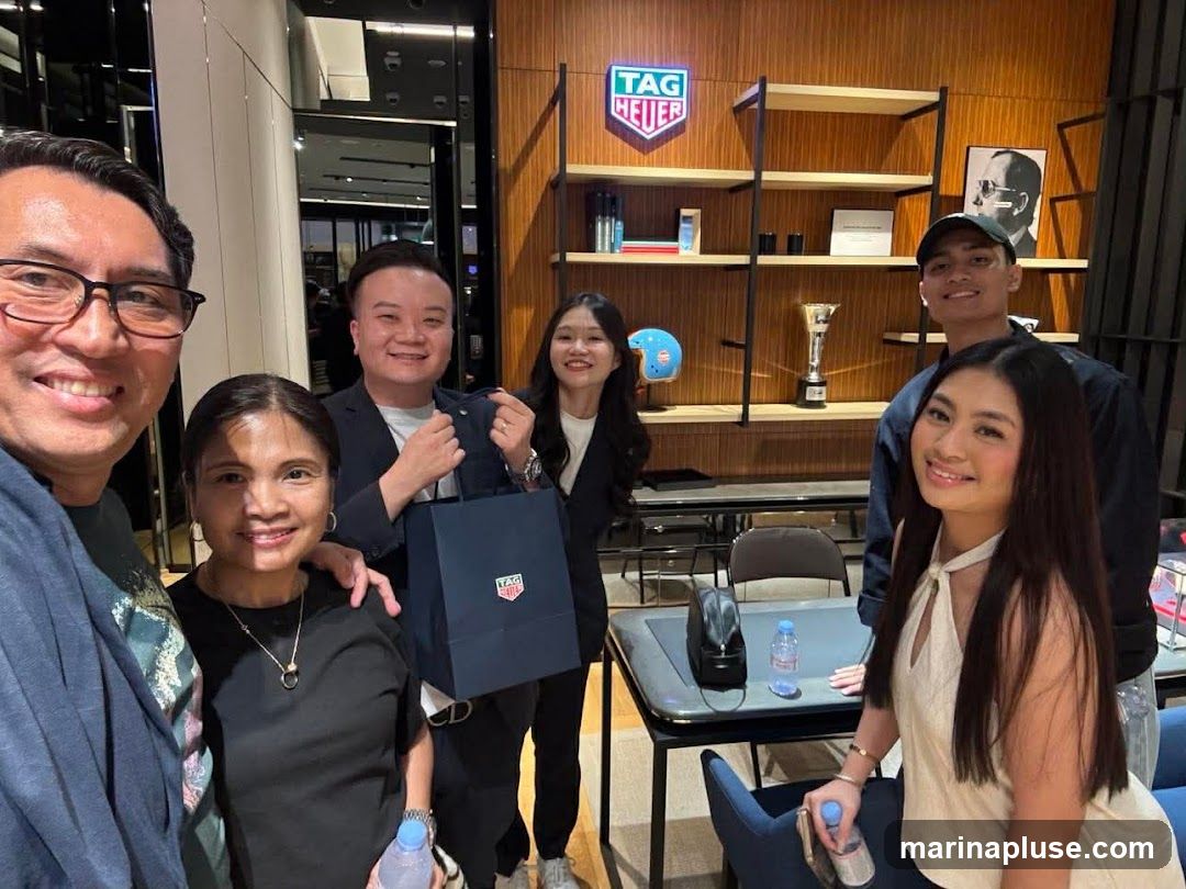 A group of five people are posing for a photo inside a store. The store appears to be a luxury watch retailer, with a prominent TAG Heuer logo displayed on the wall. The shelves behind them are stocked with various items, including what appear to be watches and accessories. One person is holding a TAG Heuer shopping bag.