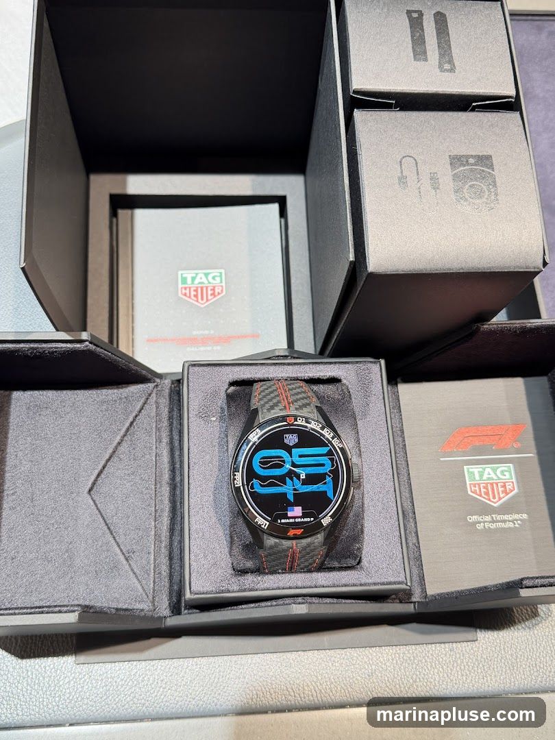 A TAG Heuer smartwatch with a black and red checkered strap is displayed in its open box. The watch face shows a digital display with racing-themed graphics. The box also contains documentation and accessories.
