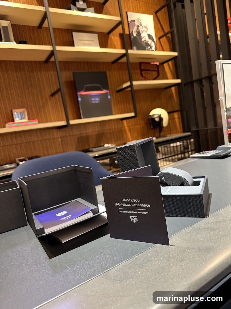 A display inside a TAG Heuer store featuring product packaging, informational booklets, and decorative items on shelves. The items include a watch box, a booklet titled "Unlock your TAG Heuer experience", and a framed picture of a man. Shelves display books, a framed image of a car, and a helmet.