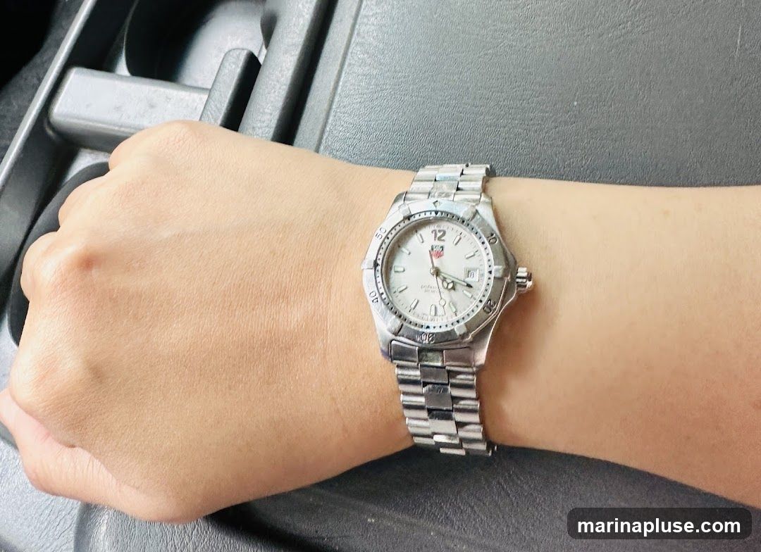 A close-up shot of a person's wrist wearing a silver watch. The watch has a white face with silver hour markers and hands. The brand logo is visible on the face. The watch band is also silver and made of links. The background appears to be the interior of a car.