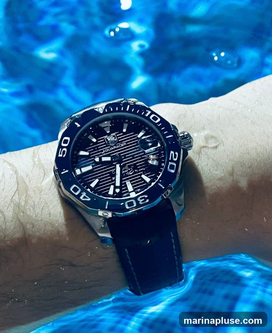 A close-up shot of a TAG Heuer Aquaracer watch on a wrist submerged in blue water. The watch has a black dial with white markers and a blue bezel. The water is rippling, creating a dynamic background.
