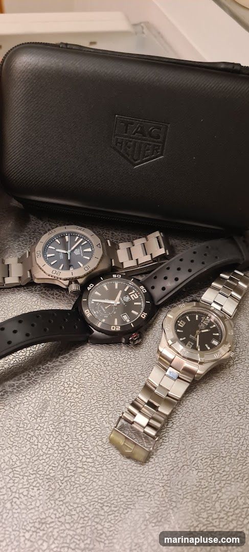 A black TAG Heuer watch case is open, revealing three TAG Heuer watches. One watch has a silver metal band and a dark blue face. Another watch has a black rubber strap and a black face with white numerals. The third watch has a silver metal band and a black face with white numerals.