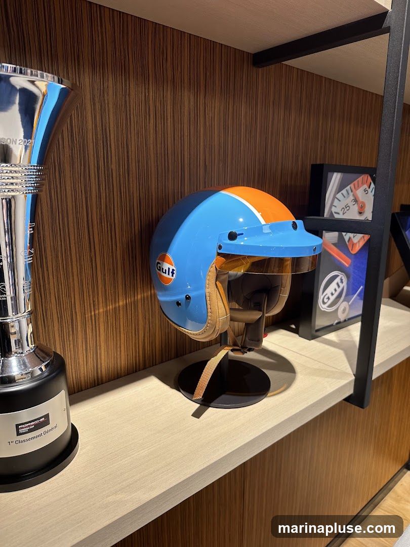 A blue and orange Gulf racing helmet is displayed on a stand on a shelf. To the left is a silver trophy with "1er Classement Général" and "PORSCHE" visible. To the right, a framed picture with a gauge and the Gulf logo is visible. The background is a wood-paneled wall.