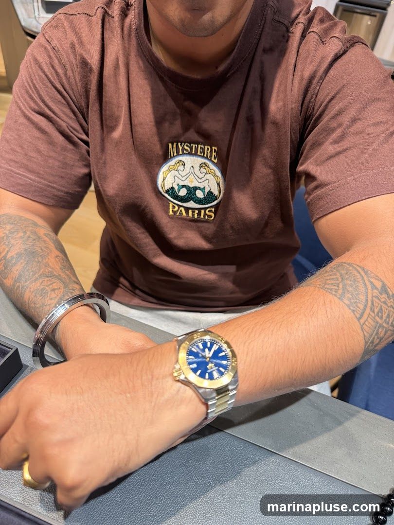 A man wearing a brown t-shirt with an embroidered logo that says "MYSTERE PARIS" and features two mermaids. He is also wearing a silver bracelet on his left wrist and a gold and silver watch on his right wrist. He has tattoos on both arms. The background appears to be an indoor setting, possibly a store.