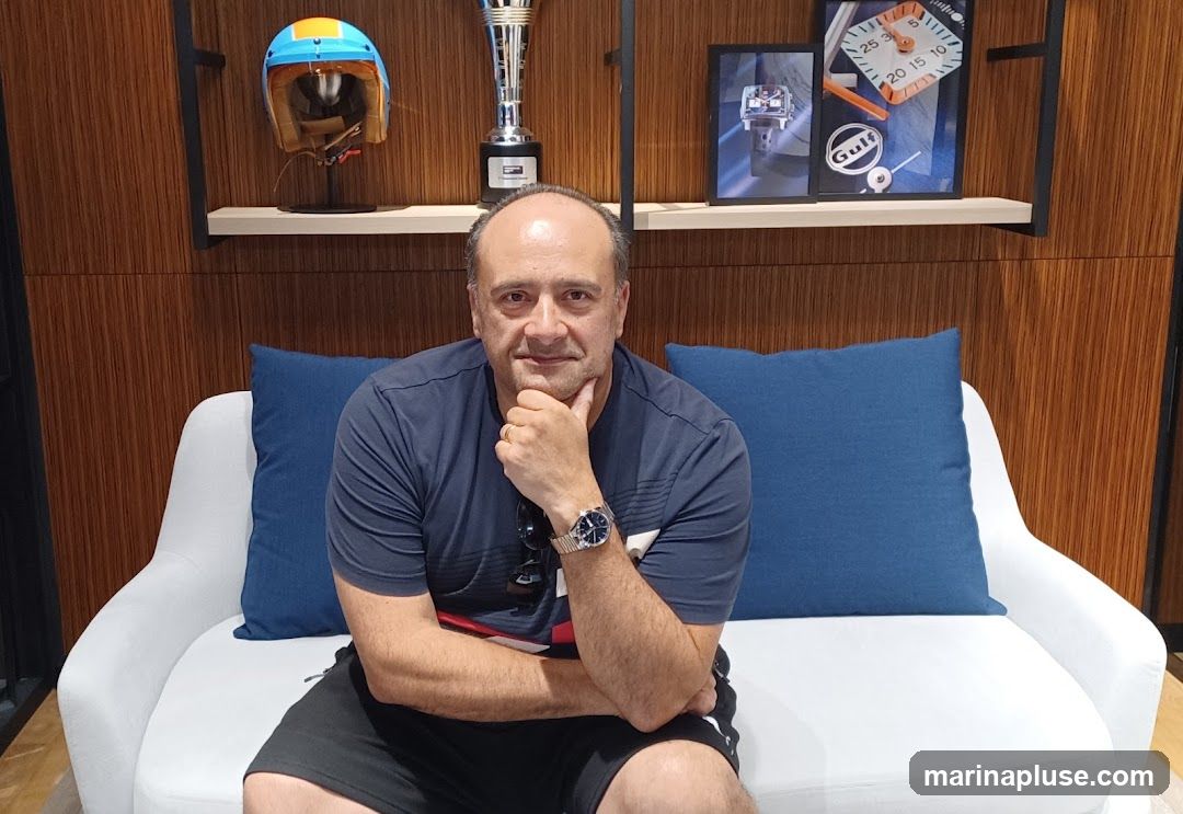 A man is sitting on a white couch with blue cushions. He is wearing a dark blue t-shirt and black shorts. He has a watch on his left wrist. Behind him, on a shelf, are a racing helmet, a trophy, and framed pictures of watches and the Gulf logo. The wall behind the shelf is made of wood panels.
