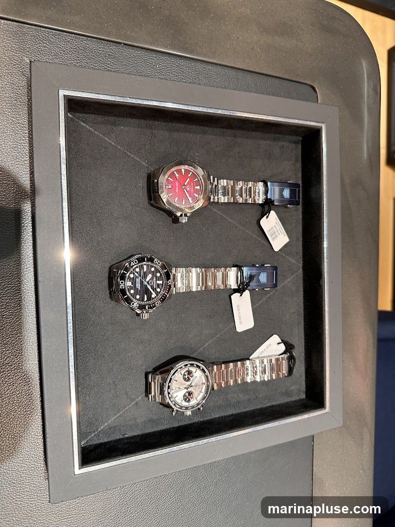 A display case holds three luxury watches with metal bands. The top watch has a red dial, the middle watch has a black dial with white markers, and the bottom watch has a silver dial with sub-dials. Each watch has a price tag attached to its band.