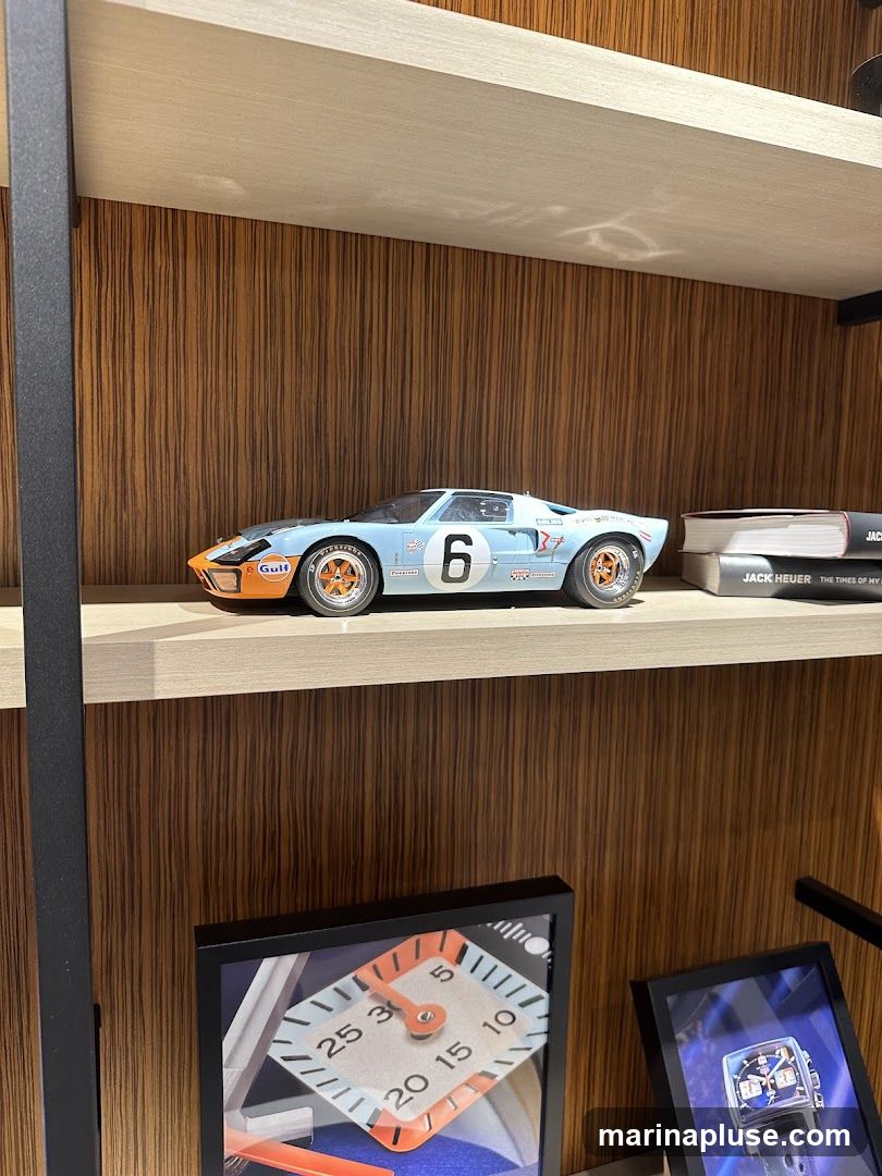 A model of a blue and orange Gulf racing car with the number 6 is displayed on a shelf. To the right of the car are two books, one titled 'JACK HEUER THE TIMES OF MY'. Below the shelf, two framed pictures are visible, one showing a watch face with orange accents and numbers, and another showing a blue square watch. The background of the shelf is a wood-grain pattern.