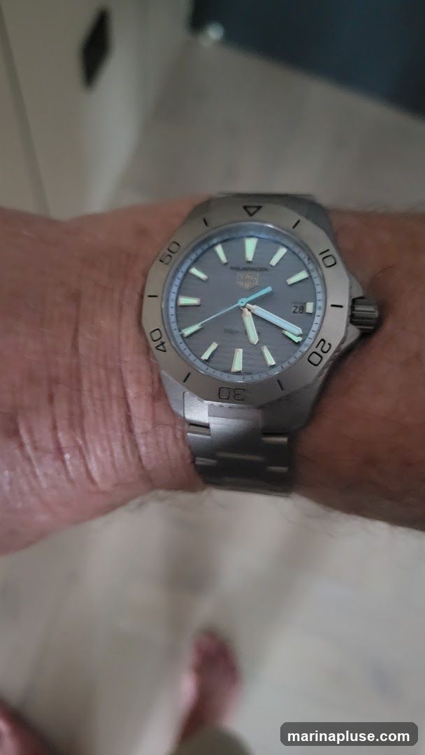 A close-up shot of a person's wrist wearing a silver watch. The watch has a grey dial with luminous hour markers and hands. The bezel is marked with numbers from 0 to 60. The watch band is also silver. The background is blurred, showing a hint of a tiled floor and a person's feet.