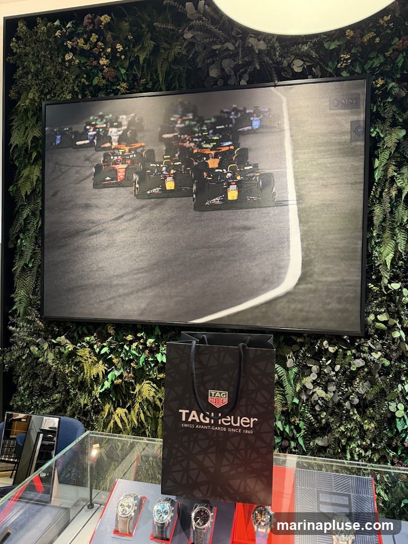 A display featuring a framed image of Formula 1 race cars, a TAG Heuer shopping bag, and several watches in a display case. The background is decorated with artificial greenery.