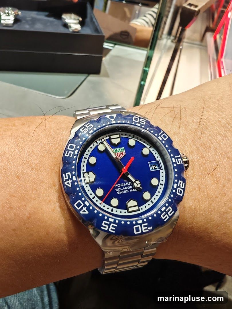 A close-up shot of a person's wrist wearing a blue TAG Heuer Formula 1 Solar-graph watch. The watch has a stainless steel band and bezel with blue markings. In the background, other watches are displayed in a dark case.