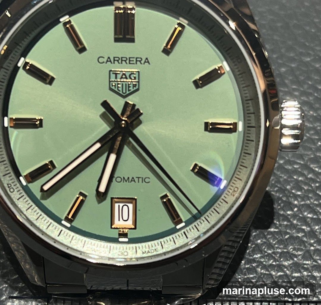 A close-up shot of a light green Tag Heuer Carrera watch face. The watch has gold-toned hour markers and hands. The brand logo and "CARRERA" are visible at the top of the dial, with "AUTOMATIC" below the center. A date window shows the number 10. The watch is on a metal bracelet.