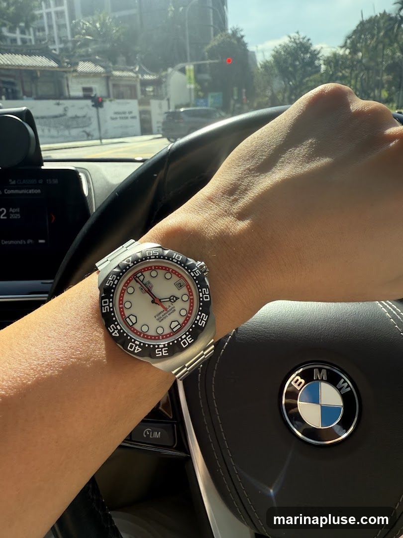 A person's arm wearing a silver watch with a white face and red accents is visible, resting on the steering wheel of a car. The BMW logo is prominent on the steering wheel. The car's dashboard display shows the date April 2025 and the time 15:58. The background shows a street scene with buildings and trees, under a bright sky.