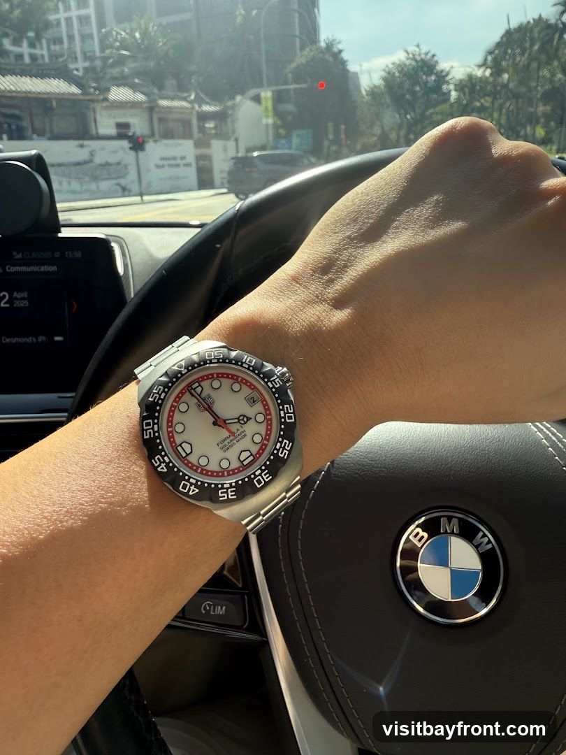 A person's arm wearing a silver watch with a white face and red accents is visible, resting on the steering wheel of a car. The BMW logo is prominent on the steering wheel. The car's dashboard display shows the date April 2025 and the time 15:58. The background shows a street scene with buildings and trees, under a bright sky.