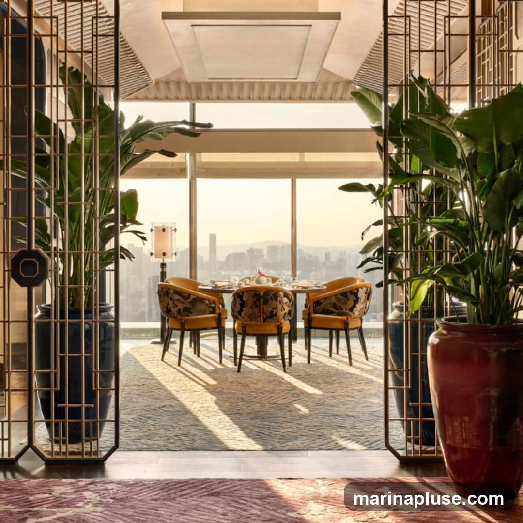 A beautifully decorated dining area with a table set for a meal, overlooking a cityscape at sunset. The room features ornate chairs with patterned upholstery, large potted plants in decorative vases, and intricate metal screens. Sunlight streams through the large windows, casting long shadows across the patterned carpet.