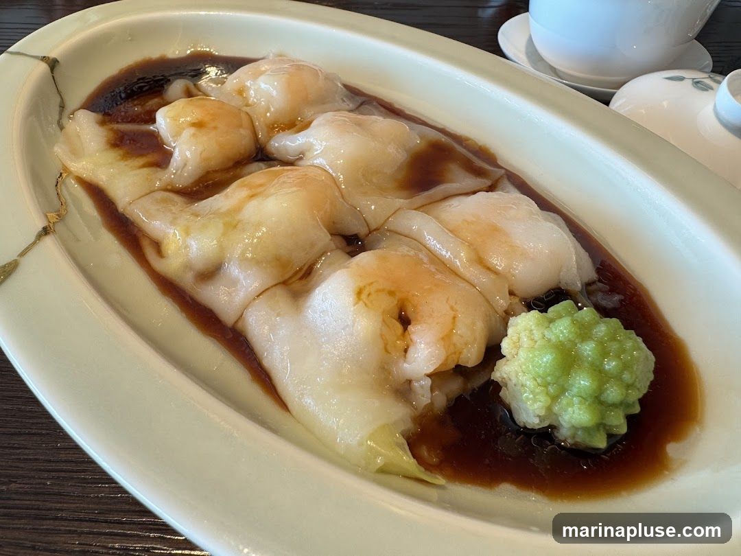 A close-up shot of shrimp rice noodle rolls served in a light-colored oval dish. The rolls are filled with plump shrimp and coated in a dark, savory sauce. A small mound of bright green garnish, possibly wasabi or a vegetable puree, sits to the side. The dish is presented on a dark wooden surface. In the background, a white teacup and saucer are visible.