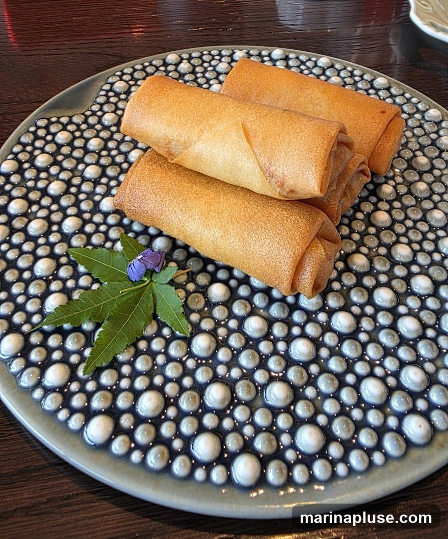 Three golden-brown spring rolls are arranged on a decorative plate with a textured pattern of raised white and grey circles. A green leaf and a small purple flower garnish the plate.