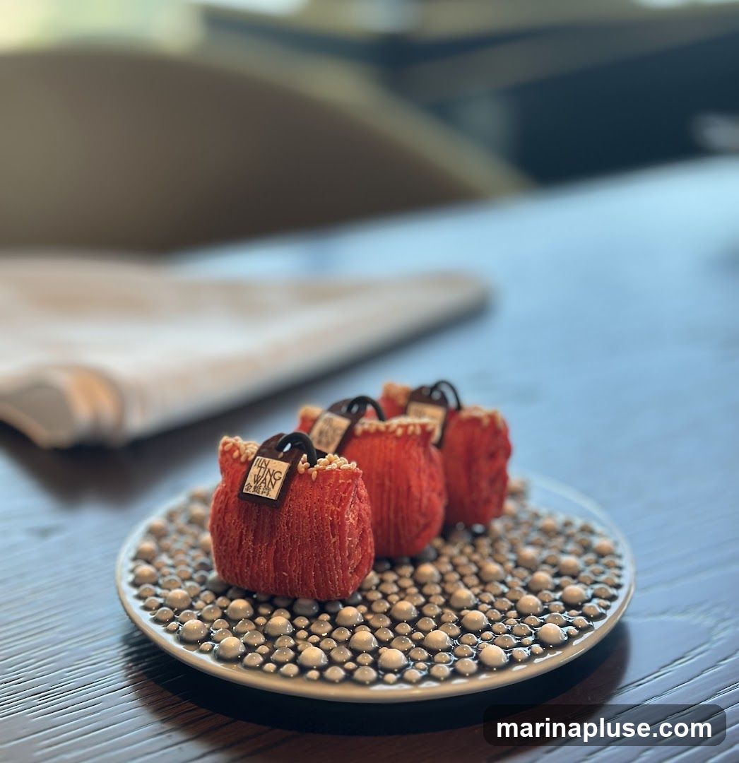 Three small, red, purse-shaped desserts are arranged on a decorative plate filled with small, pearl-like spheres. Each dessert has a dark chocolate handle and a small tag with text that appears to be a logo. The plate is on a dark wooden table, with a blurred background suggesting an indoor setting, possibly a restaurant.