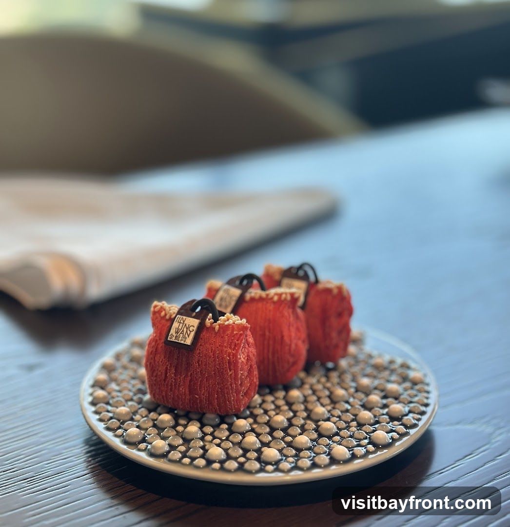 Three small, red, purse-shaped desserts are arranged on a decorative plate filled with small, pearl-like spheres. Each dessert has a dark chocolate handle and a small tag with text that appears to be a logo. The plate is on a dark wooden table, with a blurred background suggesting an indoor setting, possibly a restaurant.