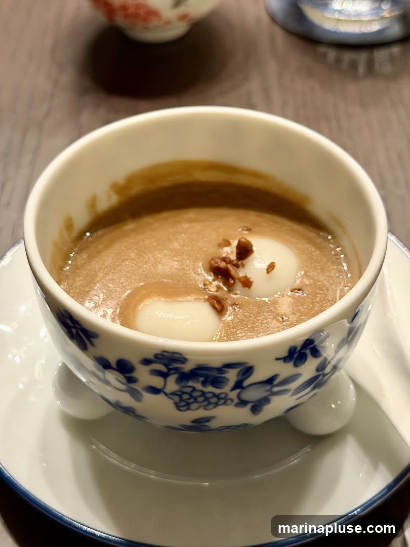 A close-up shot of a dessert in a decorative bowl. The dessert appears to be a creamy, light brown liquid with two white mochi balls floating in it. The mochi balls are topped with small, dark brown crumbles, possibly nuts or toasted grains. The bowl itself is white with a blue floral and grape vine pattern around its base and rests on a matching saucer. In the blurred background, another small bowl with red and white floral patterns is visible.