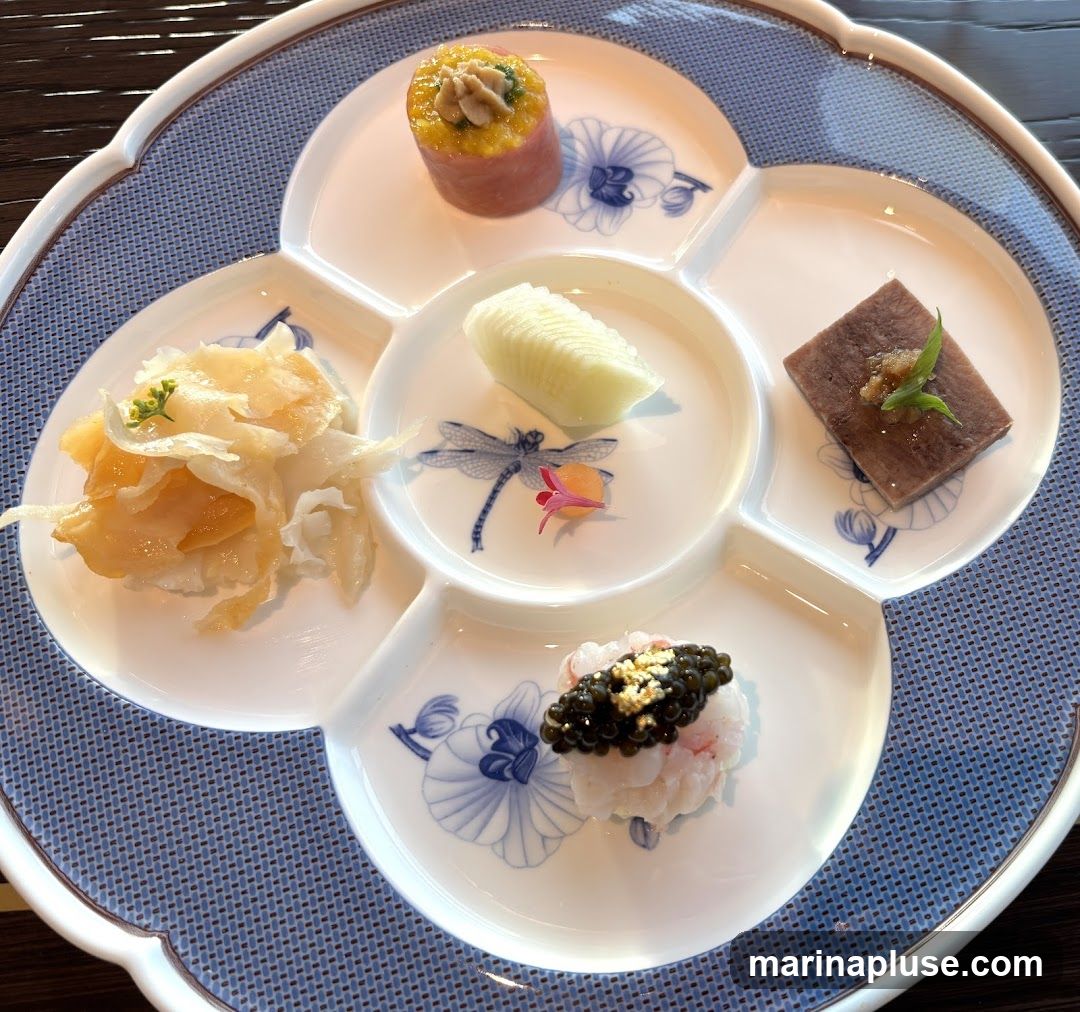 A multi-compartment white platter with blue floral and dragonfly motifs holds several small appetizers. The appetizers include a cylindrical item topped with a dollop of yellow paste and wrapped in pink meat, a piece of thinly sliced white vegetable, a square of dark meat with a garnish, a pile of translucent, layered food, and a small mound of white seafood topped with black caviar and gold flakes. The platter is set on a blue and white patterned placemat.