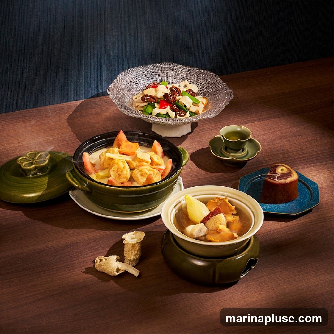 A spread of various dishes and a dessert are arranged on a wooden table. In the foreground, a steaming pot contains a stew with tofu, carrots, and tomatoes. To its right, a smaller bowl on a warmer holds a clear broth with ingredients like mushrooms and what appears to be meat. Behind these, a glass bowl contains a salad with lotus root, peppers, and nuts. A small teacup and saucer are placed near the salad, and a slice of cake with a circular topping sits on a blue hexagonal plate. Dried mushrooms are scattered on the table in the foreground.