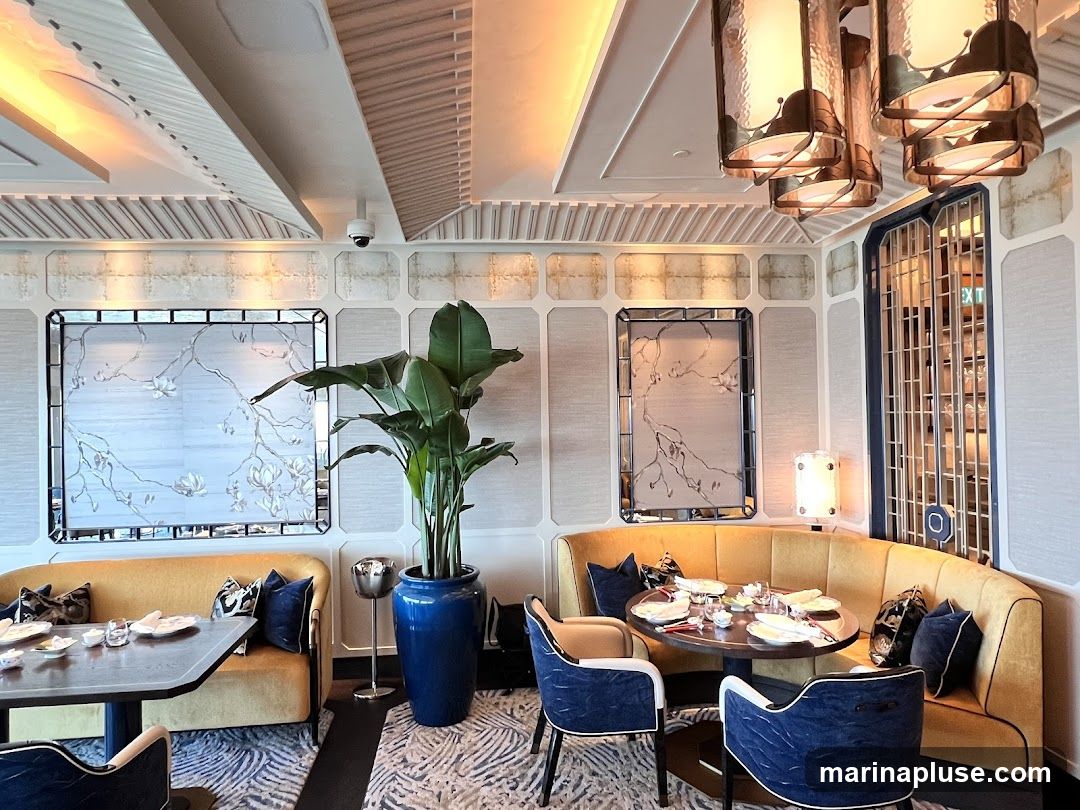 A well-lit restaurant dining area with elegant decor. Yellow banquette seating curves around a dark wood table set for a meal. Blue upholstered chairs with white accents are placed around the table. A large potted plant in a blue ceramic pot stands near the seating. Decorative framed artwork with a floral motif adorns the wall. Ornate pendant lights hang from the ceiling. A metal ice bucket is placed on a stand. The flooring features a patterned carpet.