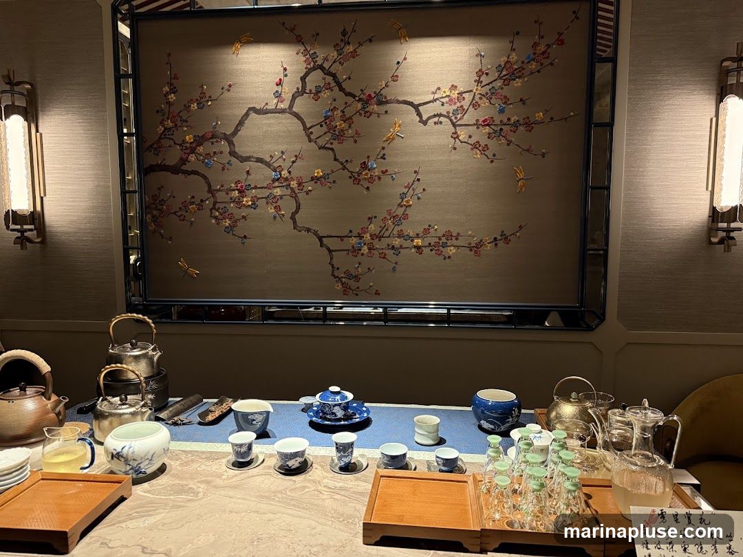 A table is set for a tea ceremony with various teapots, cups, and carafes. A decorative screen with a floral and dragonfly motif hangs on the wall behind the table. Two wall sconces illuminate the scene.